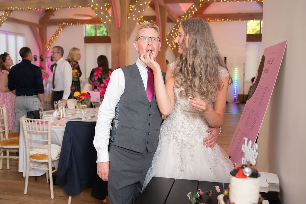Julie Fox and her husband laughing as they taste their wedding cake in the oak barn at Glebe Barn