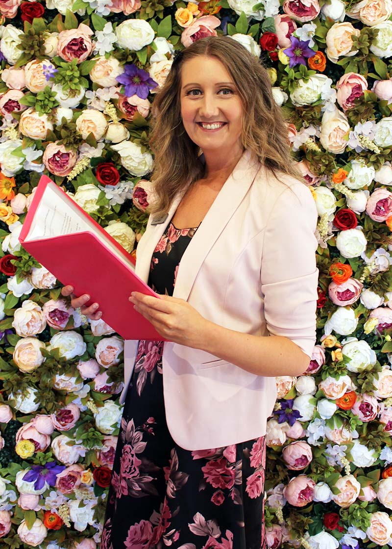 Julie Fox, wedding celebrant in Exeter, holding her ceremony folder in front of a flower wall